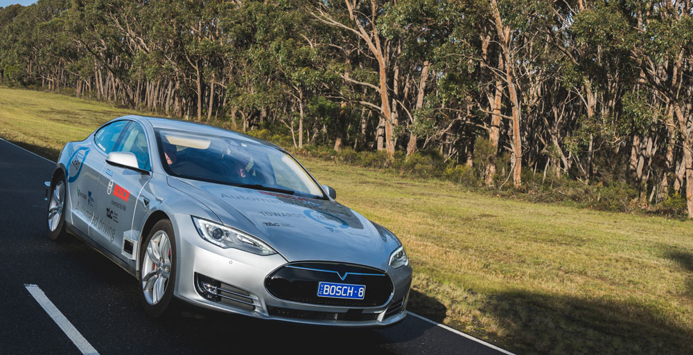 An image of a silver car with sponsors logos travelling on a road in a rural environment.